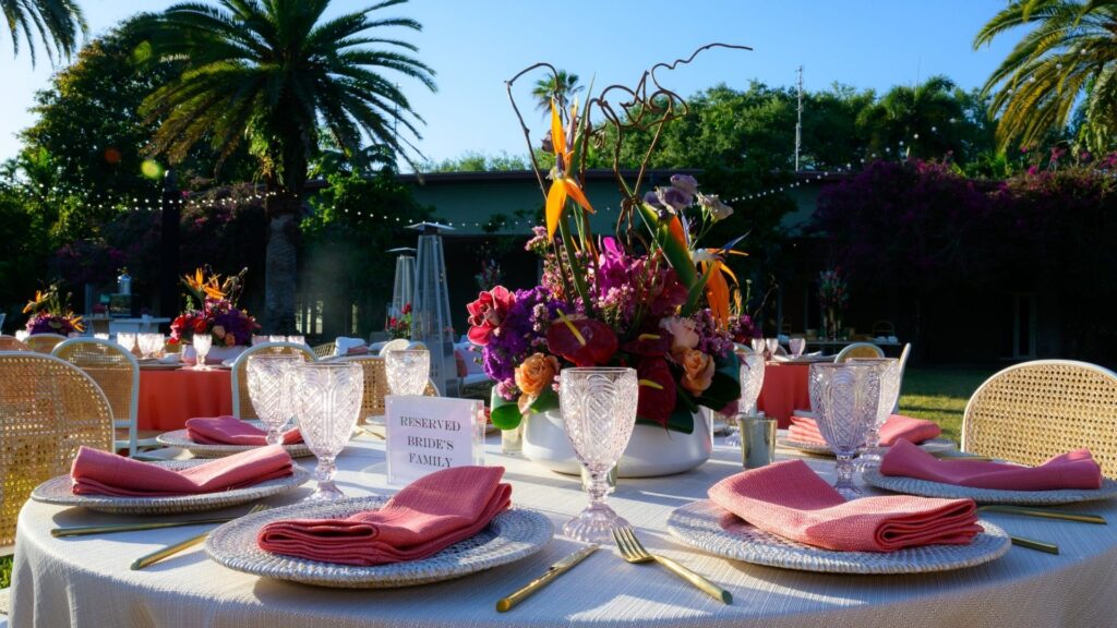 Elegant outdoor wedding reception table set with pink napkins, crystal glasses, and a vibrant floral centerpiece under palm trees at sunset.