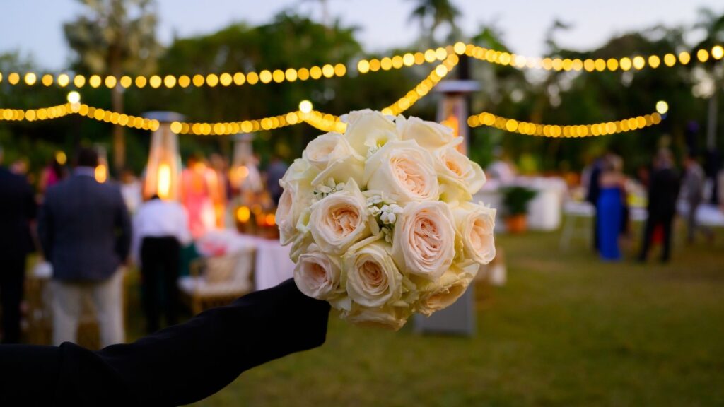 A hand holding a bouquet of white roses with warm string lights and wedding guests blurred in the background.