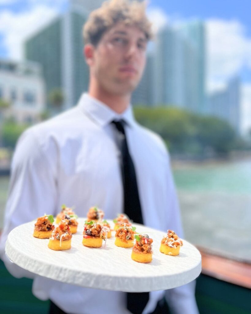 UCD server holding tray of elegant canapés on yacht with Miami skyline backdrop