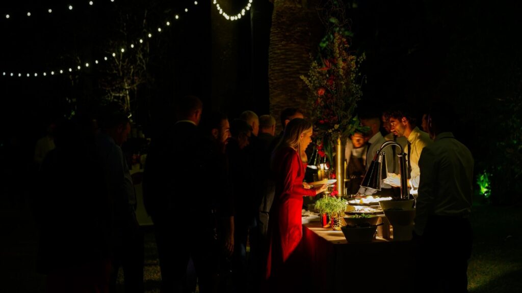 Guests in evening attire enjoying a buffet table under string lights at a nighttime wedding.