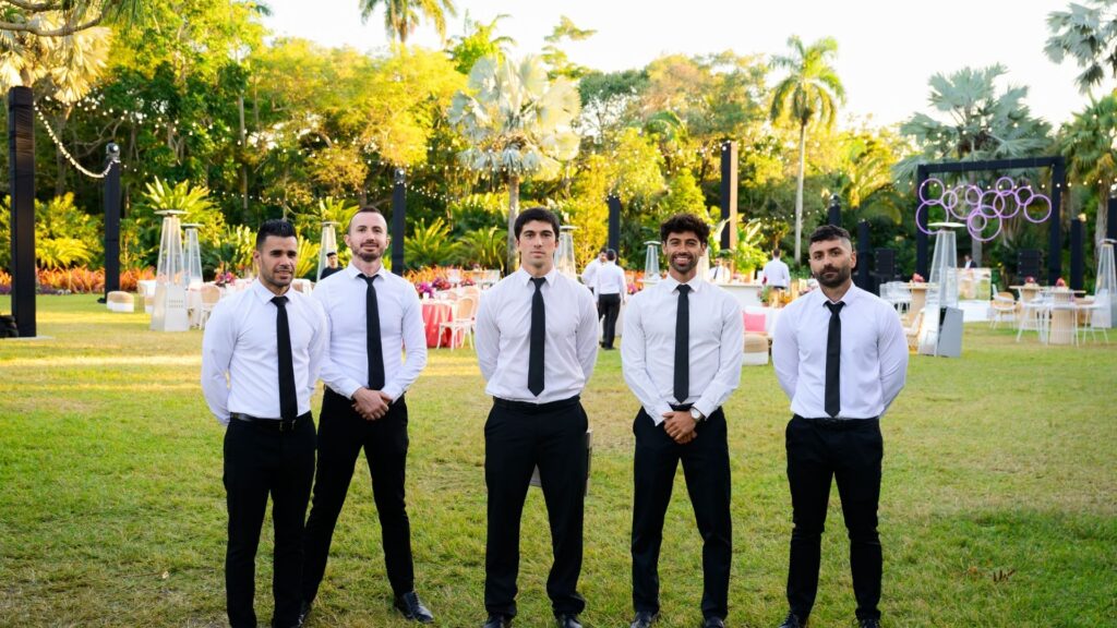 Five catering staff members in white shirts and black ties standing on a grassy lawn at an outdoor wedding reception.