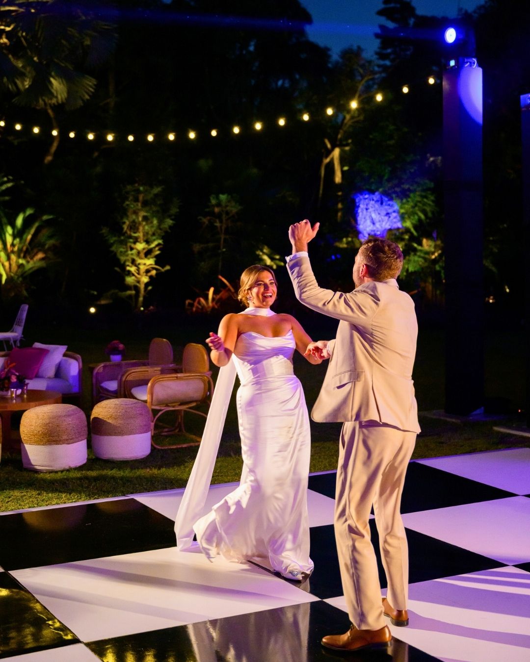 Bride and groom dancing on a black-and-white checkered dance floor outdoors at night, under string lights, surrounded by wedding lounge seating and greenery.
