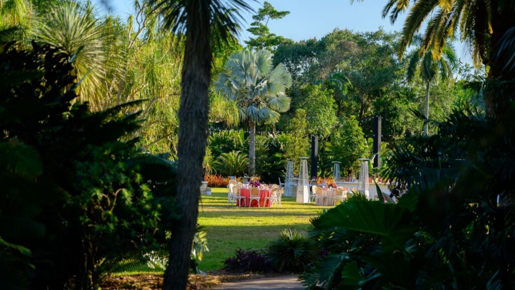 Outdoor wedding reception setup at a tropical garden venue, with round tables dressed in coral linens and surrounded by lush greenery.