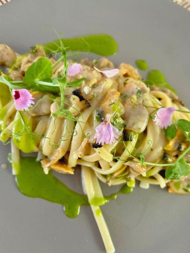 Fine dining pasta dish garnished with edible flowers and microgreens on a gray plate.