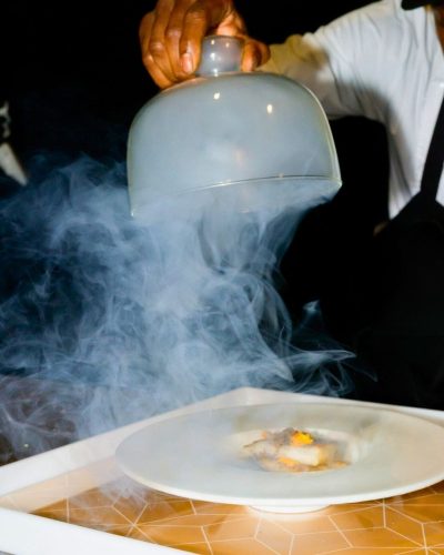 Smoke-covered reveal course Chef lifting cloche with smoke effect to reveal plated dish