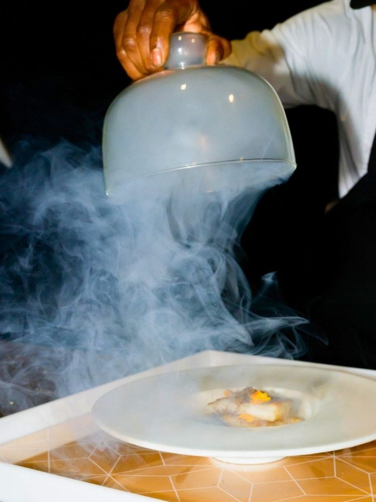 Chef lifting cloche with smoke effect to reveal plated dish
