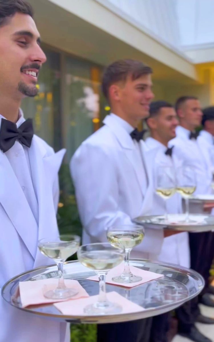 Uniformed white-glove servers holding champagne trays during an outdoor luxury event.